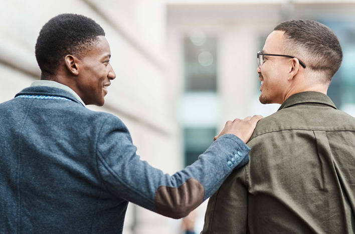 A young man placing his hand on the shoulder of another young man. The two are smiling at each other.