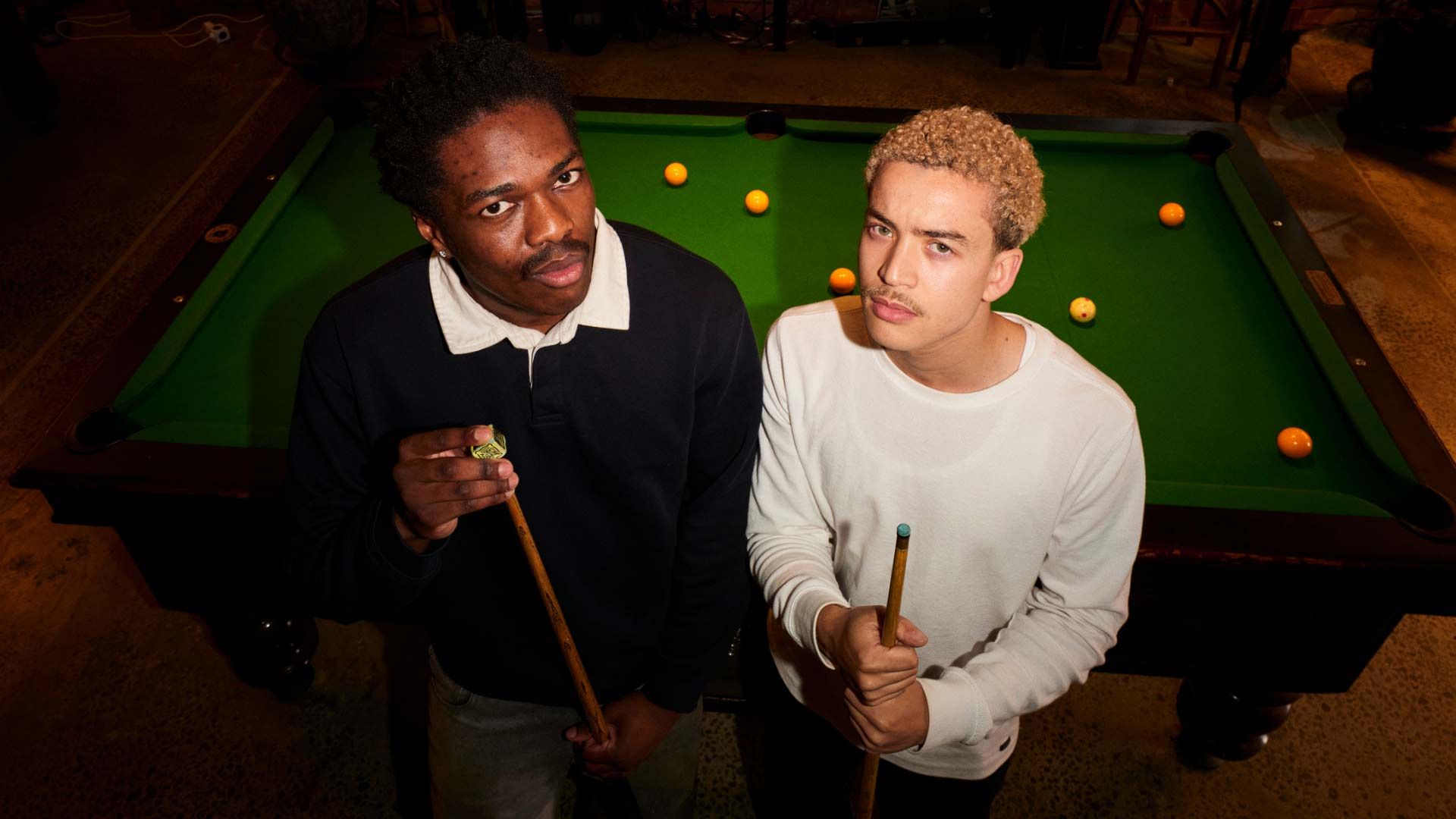 Photo of two young men playing at a pool table, looking up to camera.